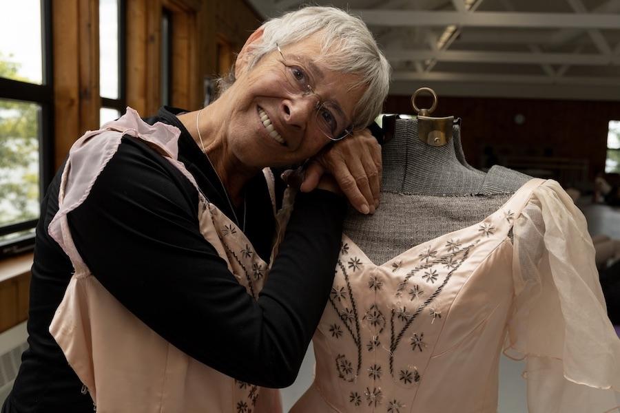 A smiling elderly woman leans on a dress frame with the dress she wore as a camper in 1962.