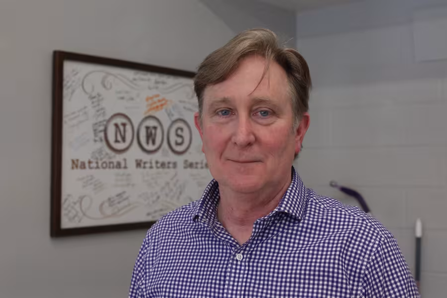 Doug Stanton smiles standing in front of a framed poster for the National Writers Series.