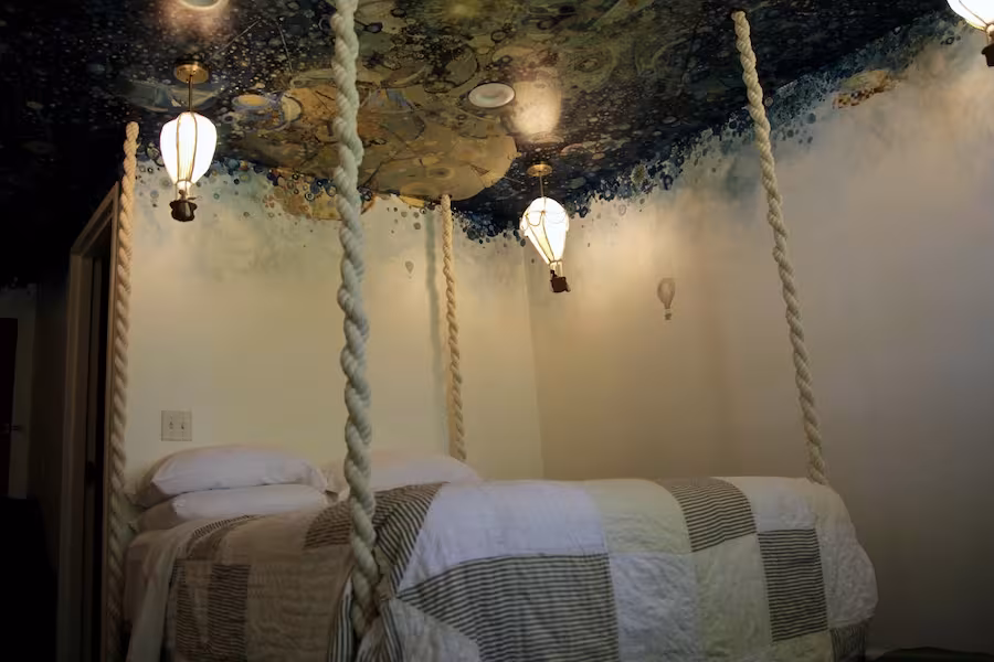A shot of a bed hung by thick ropes from the ceiling and surrounded by small hot air balloons, under a brightly-painted ceiling.