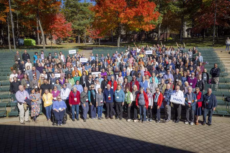 A large group of excited people gather in an outdoor auditorium, holding class year signs.