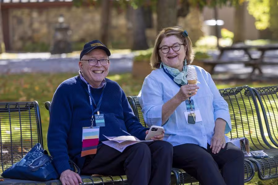 Two alumni smile while sitting on benches; one holds an ice cream cone.