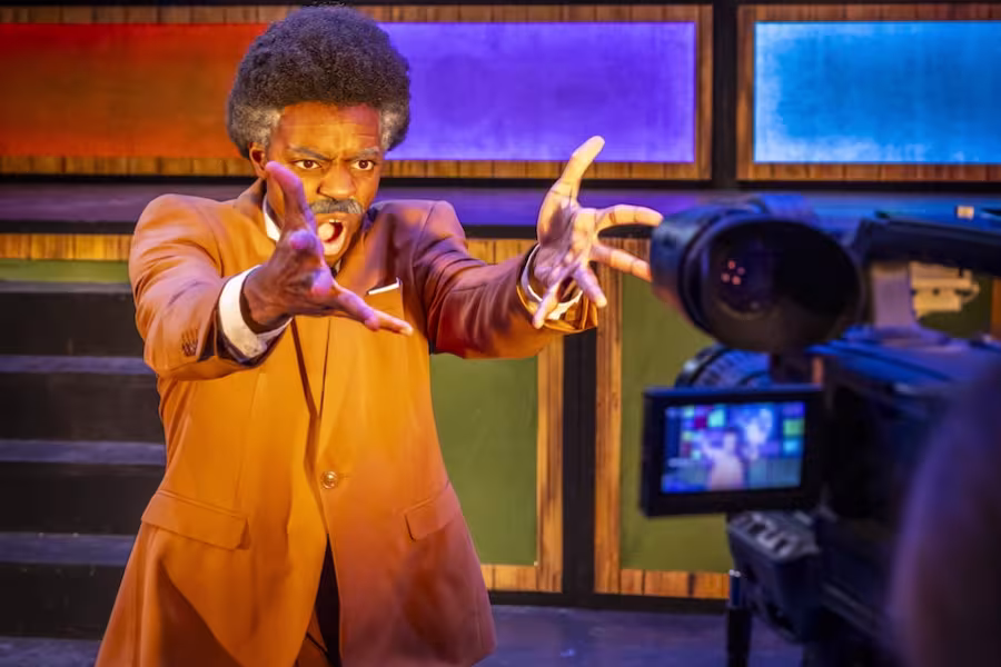 A production still from the Interlochen Arts Academy production of Network. Set in a TV studio, a man in a brown suit speaks energetically in front of a camera, colorful panel lights and a large TV screen behind him.