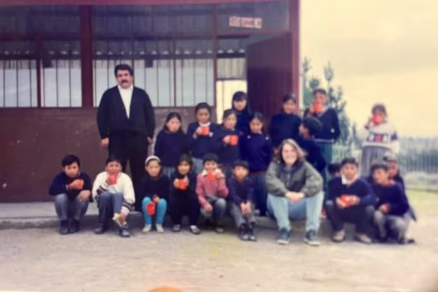 A male teacher and a class of small children sit with a woman in front of a school building.