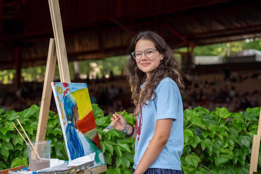 A student takes a break from painting a picture to smile at the camera.