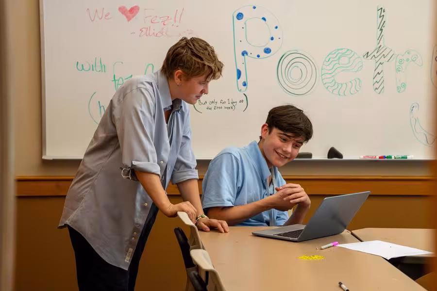 A student leans over a laptop while another student looks on.