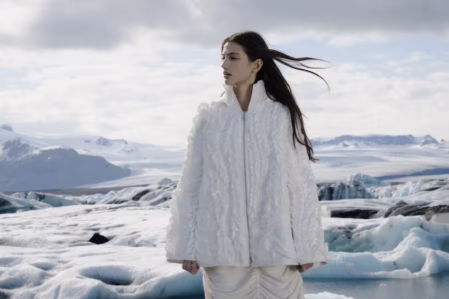 A model wearing a textured white coat poses against a wintry landscape.