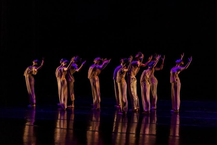 A group of dancers dressed in light brown extend their arms gracefully on a dark stage.
