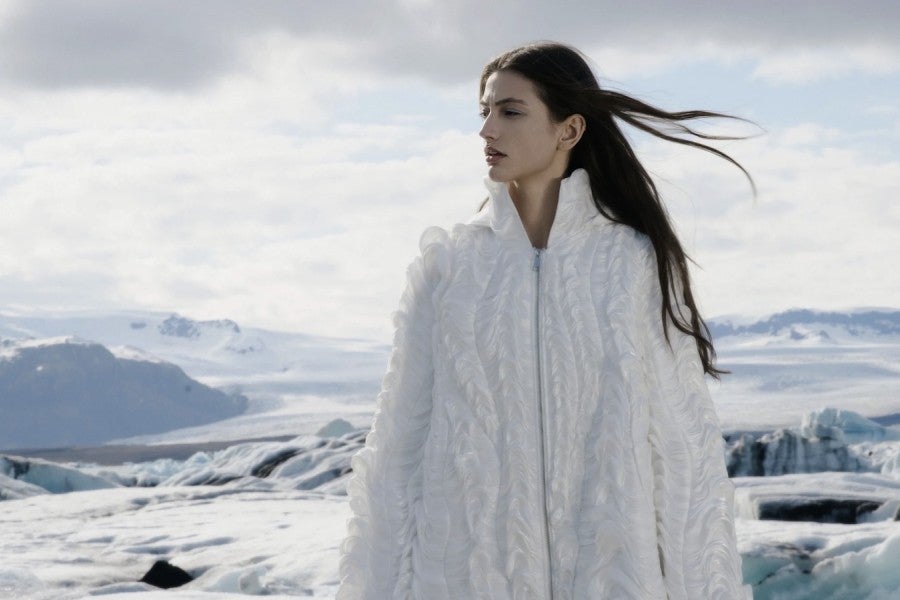 A model wearing a textured white coat poses against a wintry landscape.