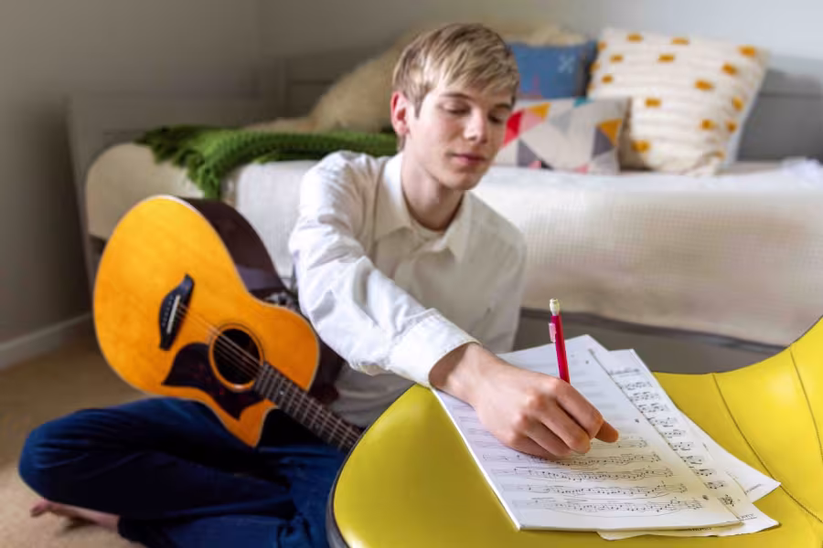A guy writes on staff paper and is holding a guitar