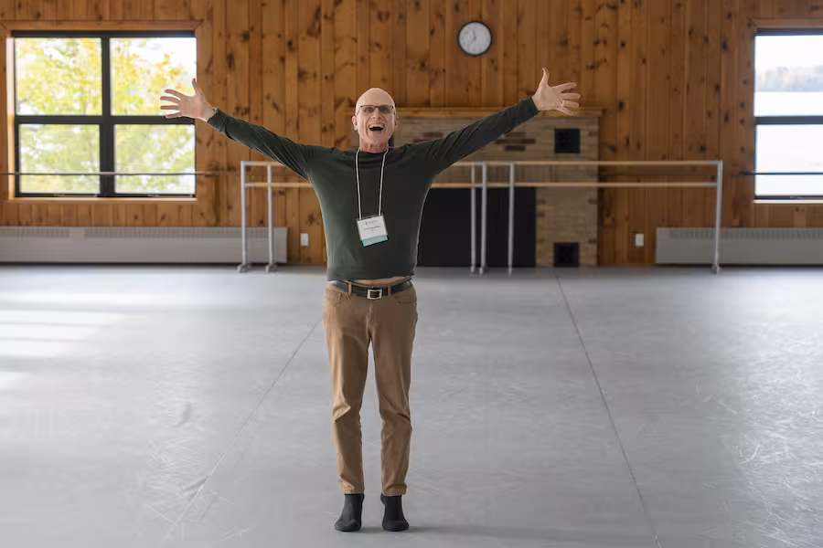 Peter Sparling stands smiling with hands outstretched in the Dance Center. 