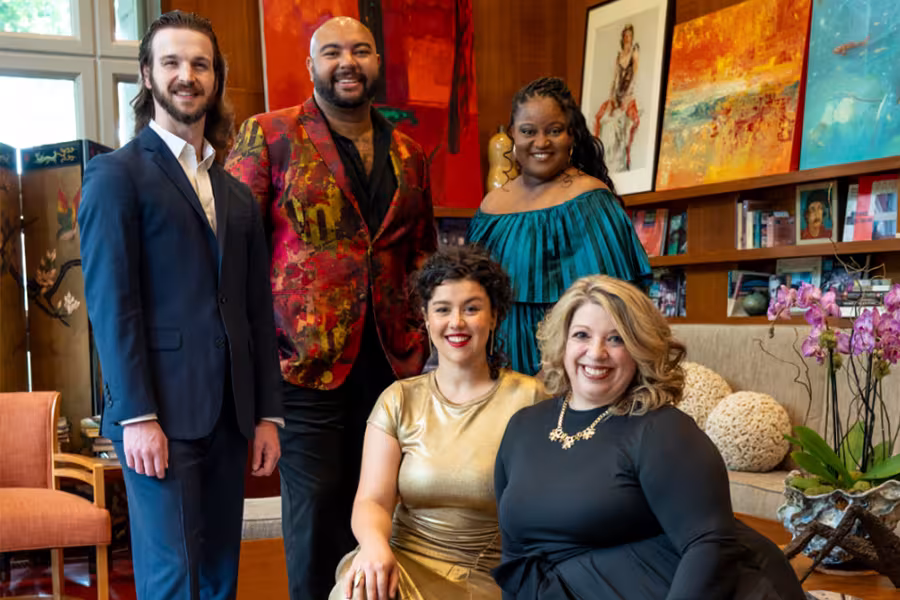 A group of five well dressed performers pose together in a warm, art filled room. Two people sit at the front, one in a gold dress and one in a black dress, while three stand behind them wearing a blue suit, a vibrant red patterned jacket, and an emerald off the shoulder dress. Colorful paintings and books line the shelves in the background.