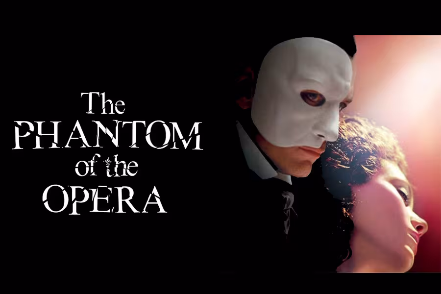 Promotional image for The Phantom of the Opera showing a masked man in a white half mask looming protectively behind a woman with dark hair, both lit by warm stage light against a dark background, with the title The Phantom of the Opera in elegant white lettering on the left.