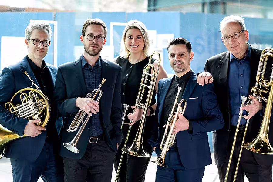Five professional brass musicians stand together indoors holding French horn, trumpet, trombone, and tuba, dressed in dark formal attire and smiling toward the camera, conveying a polished and confident ensemble presence.