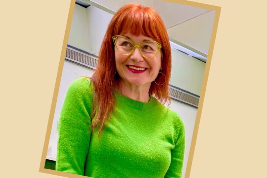 Portrait of Brooke Keesling​ with red hair and green glasses, wearing a bright green sweater, standing indoors against a light colored background.