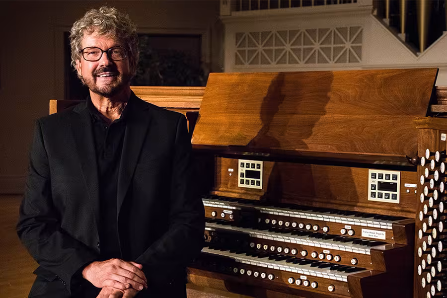 A smiling man wearing glasses and a black jacket sits beside a large pipe organ console, with multiple keyboards, rows of stop knobs, and warm wooden panels visible in a softly lit concert hall.