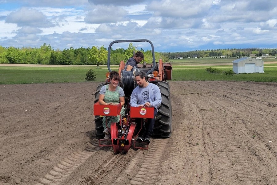 Three people sit on a potato planter as it moves through a field.
