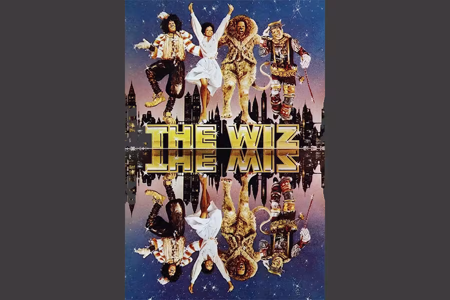 Poster for The Wiz featuring four characters — the Scarecrow, Dorothy, the Cowardly Lion, and the Tin Man — dancing joyfully against a stylized city skyline at dusk. The title “THE WIZ” appears in large gold letters at the center, mirrored below along with a reflection of the characters, creating a symmetrical effect.