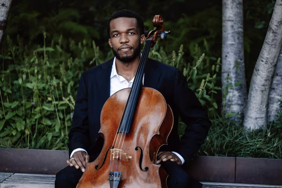 A musician wearing a suit sits with his cello against a natural backdrop.