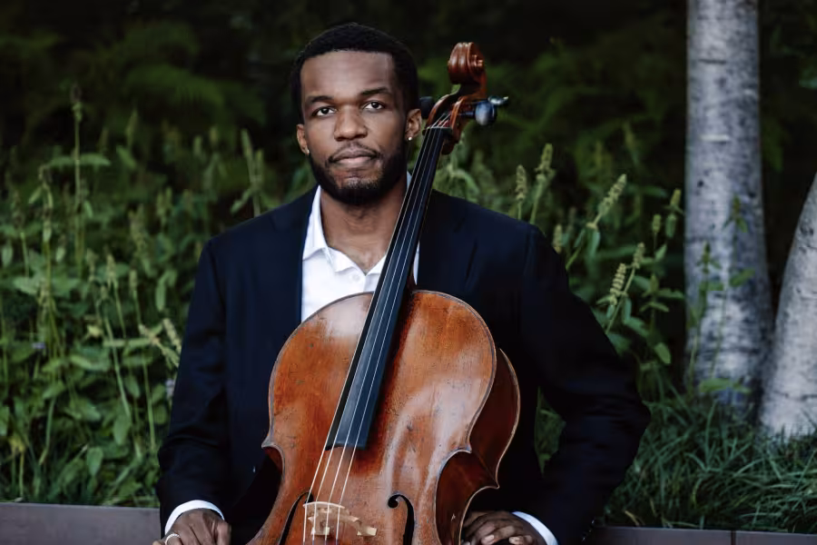 A musician wearing a suit sits with his cello against a natural backdrop.