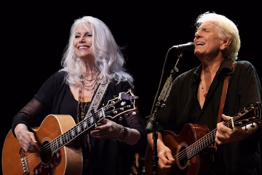 Two musicians perform on stage against a black background. A woman with long white hair smiles while playing an acoustic guitar. Beside her, a man with short white hair sings into a microphone while playing his own acoustic guitar.