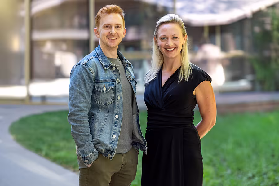 Two adults stand outdoors on a curved sidewalk with a grassy area behind them. The person on the left has short red hair and wears a denim jacket over a gray shirt and olive pants. The person on the right has long blonde hair and wears a black dress. Both are smiling and facing the camera, with blurred buildings and greenery in the background.
