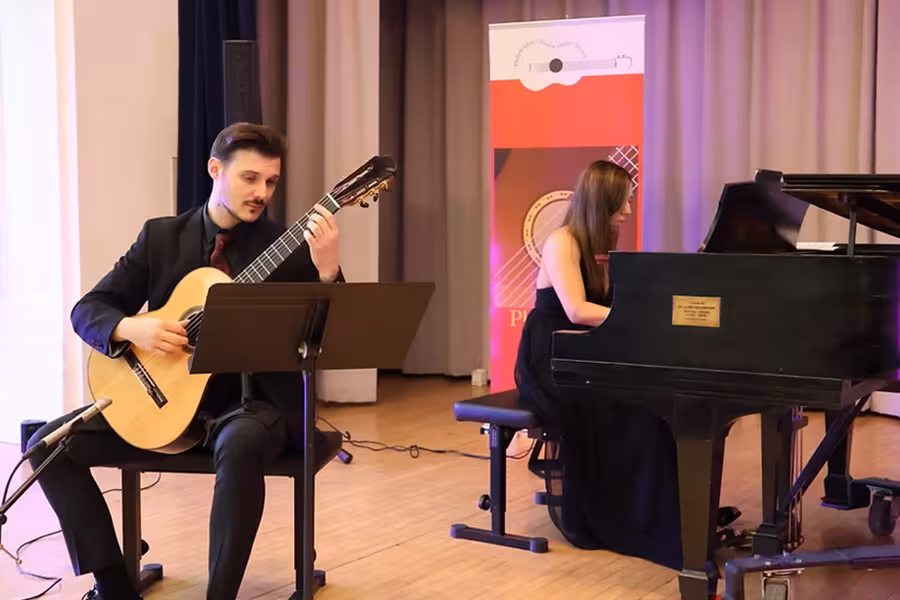 A guitarist and a pianist perform together on a small indoor stage. The guitarist, seated at left, plays a classical guitar while reading sheet music on a stand. The pianist, seated at right, plays a grand piano. Both musicians are dressed in formal black attire. A microphone is positioned near the guitar, and a colorful vertical banner stands behind them. The setting appears to be a recital or chamber music concert.
