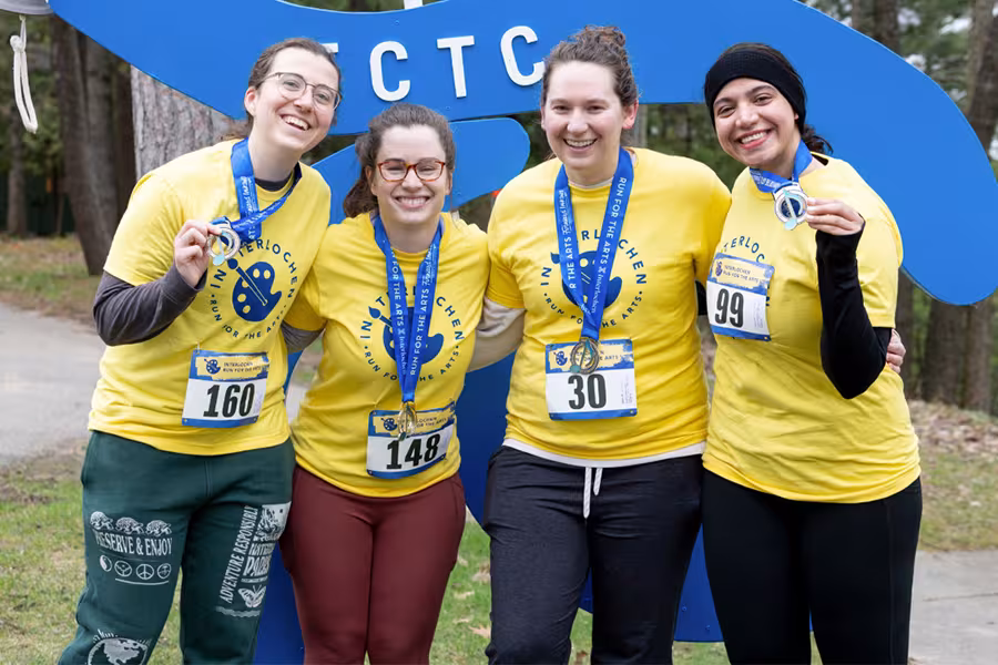 A group of four smiling runners wearing matching yellow “Interlochen Run for the Arts” shirts pose together after the race. They each hold up their medals and have race bibs pinned to their shirts. They stand in front of a large blue race logo outdoors among trees.