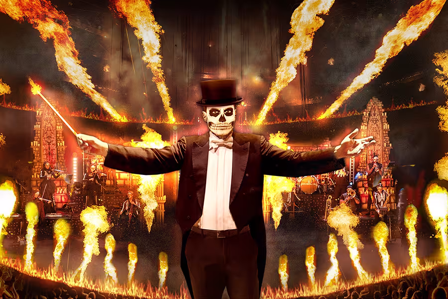 A theatrical rock concert scene featuring a conductor with a painted skull face wearing a top hat and tuxedo, standing center stage with arms outstretched and a baton in one hand. Behind him, musicians perform amid dramatic fire effects, glowing orange flames, and gothic-style set pieces. The stage is filled with intense lighting and pyrotechnics, creating a dark, high-energy atmosphere. The logo “The Rock Orchestra” appears above the conductor.