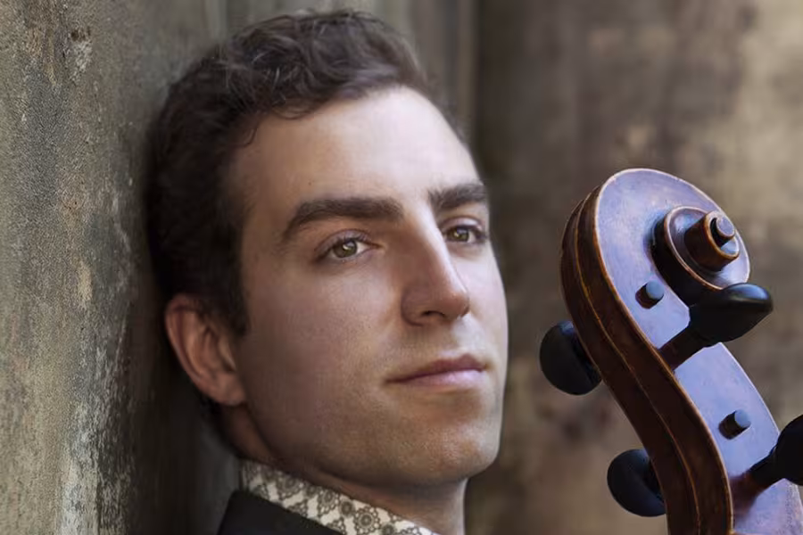 A man with short, dark curly hair leans against a textured wall while holding the scroll of a cello beside his face. He looks slightly past the camera with a calm, thoughtful expression. The light highlights his features and the warm wood tones of the cello.