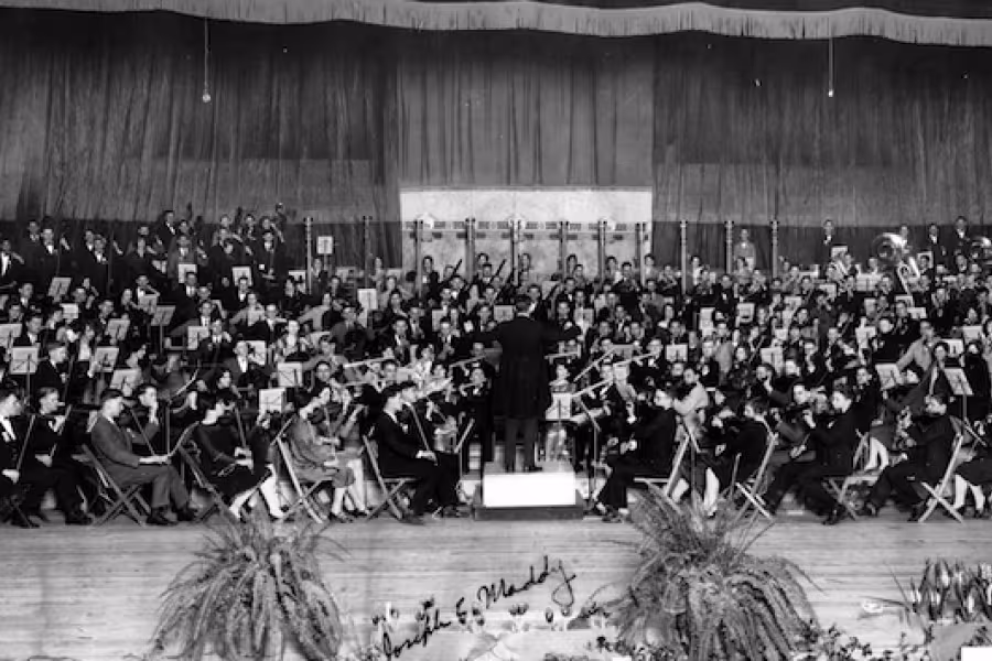 A large orchestra seated onstage with a conductor standing at the center amidst a backdrop of curtains.
