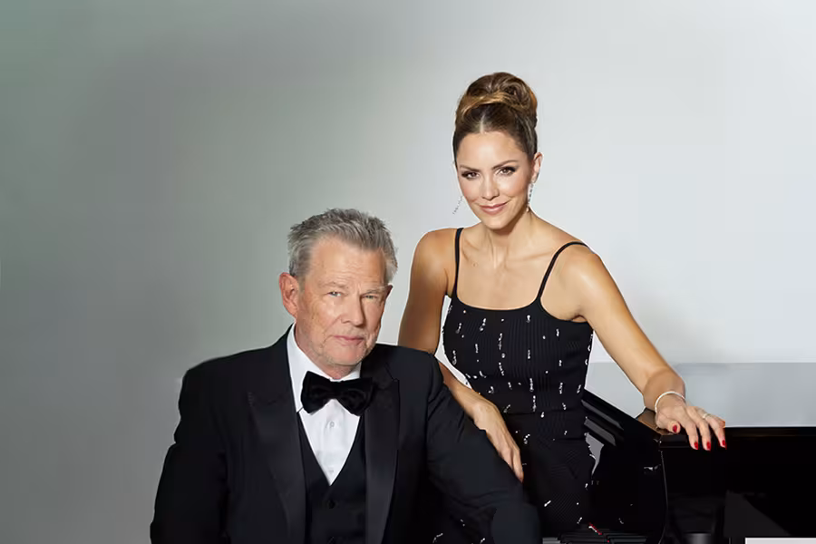 David Foster and Katharine McPhee pose together in formal attire beside a piano. Foster is seated, wearing a black tuxedo with a bow tie, while McPhee stands next to him with one hand resting on the piano, dressed in a fitted black gown with thin straps. Both look toward the camera against a simple light background.