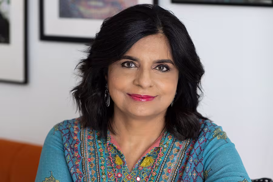 Professional headshot of a Parul Kapur with shoulder-length dark hair, wearing a teal embroidered top and earrings, smiling gently while facing the camera against a softly blurred indoor background with framed artwork.