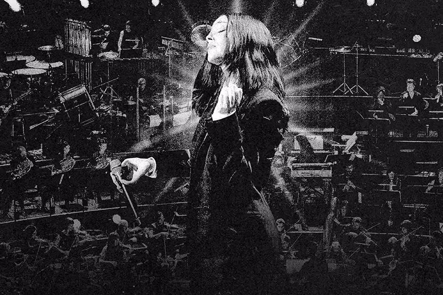 Black-and-white concert image of St. Vincent standing center stage with eyes closed, holding a conductor’s baton as beams of light radiate behind her. An orchestra and instruments are visible in the background, creating a dramatic, cinematic performance scene.