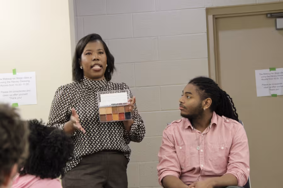 An arts instructor holds a makeup palette and gestures as she stands next to a seated student.