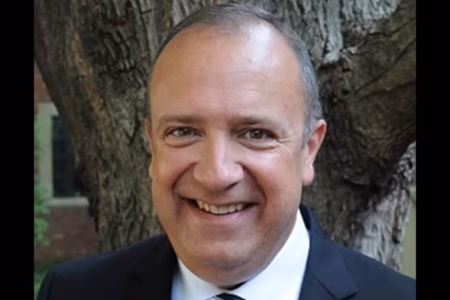 Portrait of conductor Kevin Sedatole smiling outdoors in front of a large tree, wearing a dark suit, white shirt, and tie.