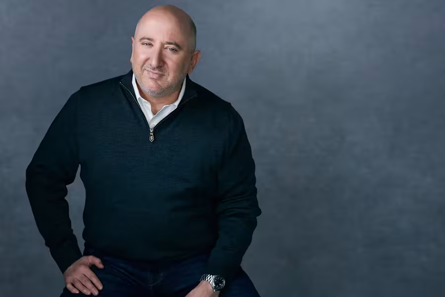 Portrait of Adam Glaser seated against a gray backdrop, wearing a dark quarter-zip sweater over a white collared shirt, with one hand resting on his knee and looking toward the camera.