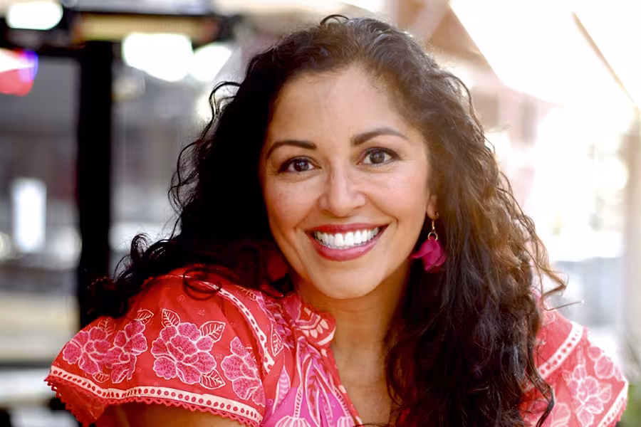 Portrait of Aimee Nezhukumatathil smiling warmly at the camera outdoors, with long curly dark hair, pink earrings, and a bright coral floral-patterned blouse. Blurred city street background with soft natural light.