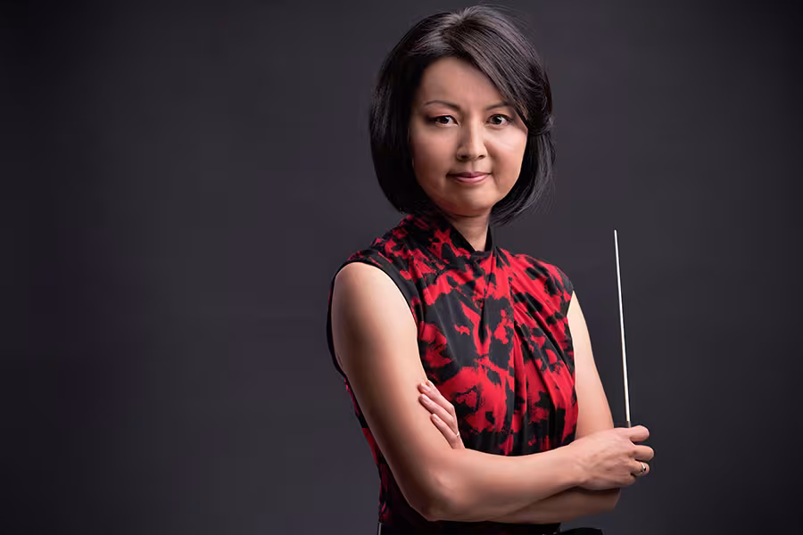 Portrait of Akiko Fujimoto with short dark hair wearing a red and black sleeveless patterned dress, standing with arms crossed and holding a conductor’s baton against a dark background.