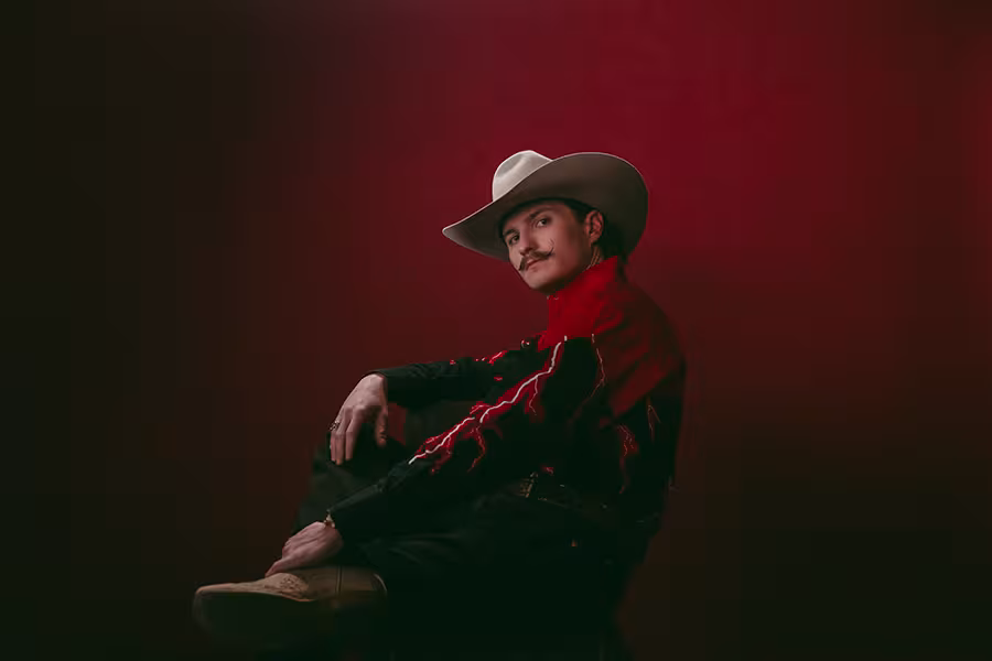 Man (Braxton Keith) wearing a white cowboy hat, red western shirt, and black pants sits in a relaxed pose against a dark red background, looking toward the camera.