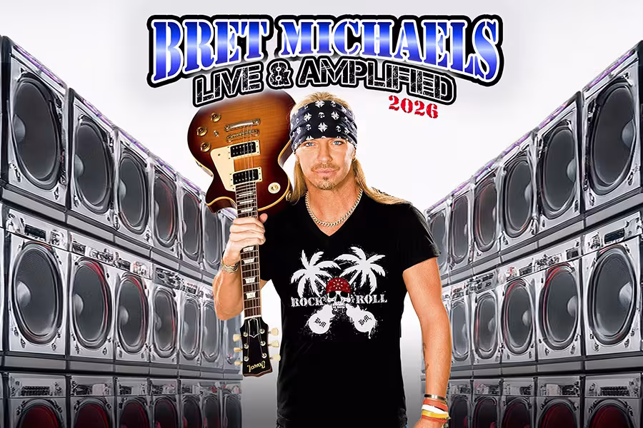 Promotional image of Bret Michaels holding an electric guitar, standing in front of a wall of stacked speakers. He wears a black T-shirt with a skull-and-palm-tree graphic, a bandana, and long blond hair. Text above reads “Bret Michaels Live & Amplified 2026.”