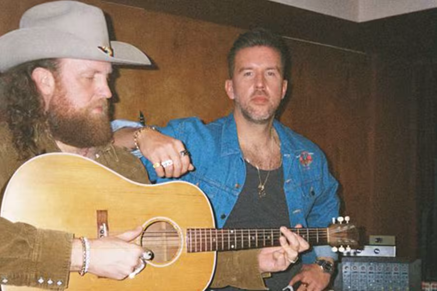 Two musicians sit indoors against a wood-paneled wall. One wears a cowboy hat and holds an acoustic guitar while looking down at the instrument. The other sits beside him in a blue jacket, facing the camera. Audio equipment is visible in the background.
