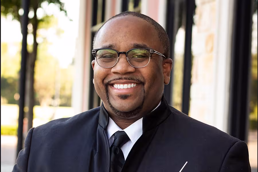 Portrait of conductor Creston Herron smiling outdoors, wearing glasses and a black formal concert jacket with white shirt and black tie, standing in front of a building with windows and trees in the background.
