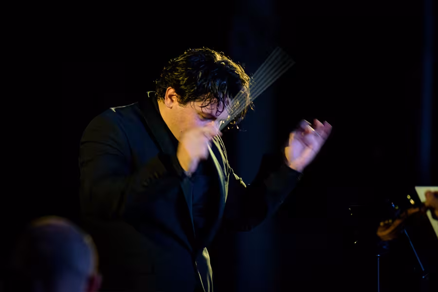 Cristian Măcelaru conducts passionately on a dimly lit stage, raising his baton with expressive motion while wearing a black suit.