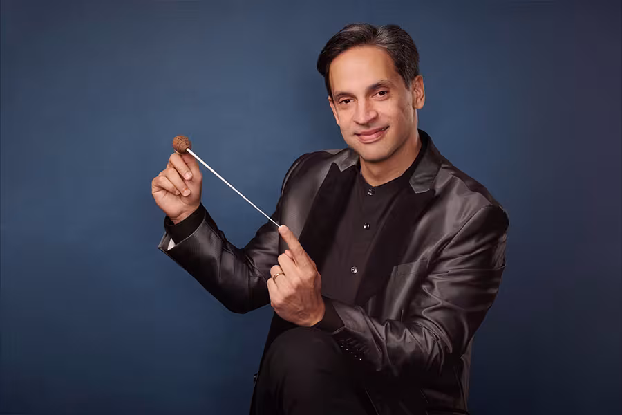 Portrait of Darwin Aquino seated against a blue background, smiling while holding a conductor’s baton. He wears a dark satin jacket over a black shirt and faces the camera in a poised, confident pose.