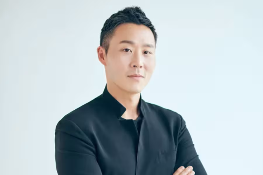 Portrait of conductor Earl Lee wearing a black jacket, standing with arms crossed against a light background.