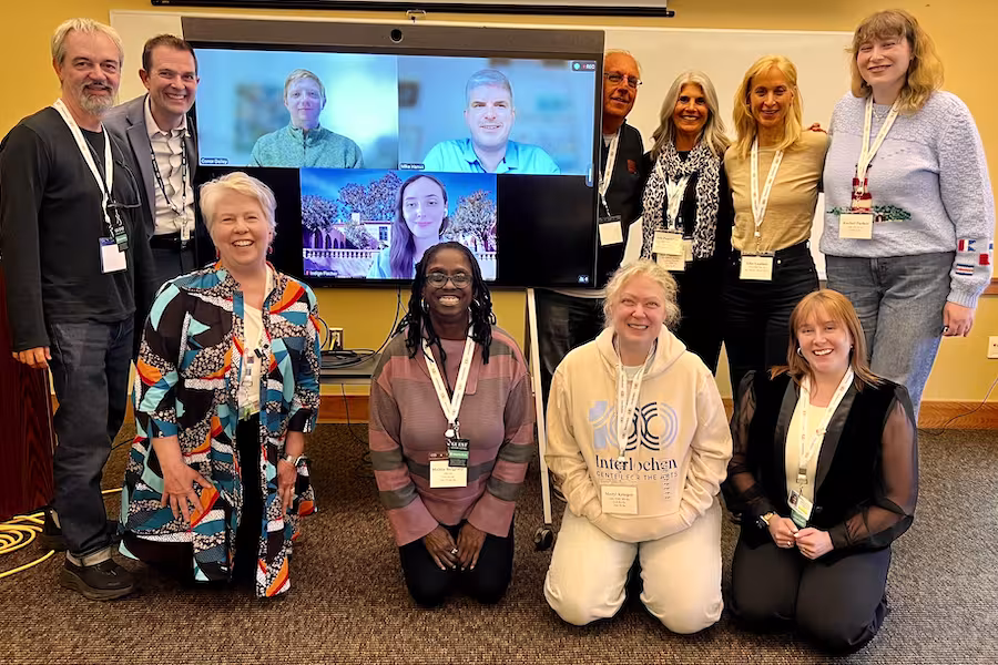 A group photo of people at a conference, with some attending virtually shown on a large monitor screen.