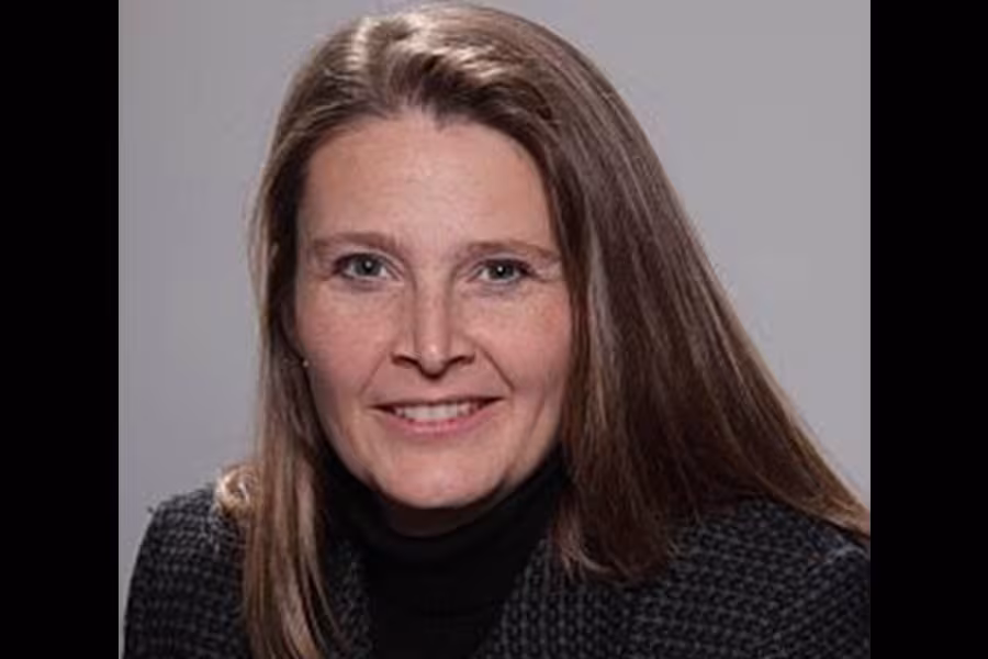 Portrait of Erica Neidlinger smiling at the camera, with long brown hair, wearing a dark blazer over a black turtleneck against a light gray background.