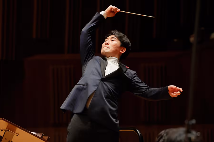 Keitaro Harada conducts energetically onstage, wearing a dark suit and white shirt, with one arm raised high holding a baton and the other extended outward against a warm wood-paneled concert hall backdrop.