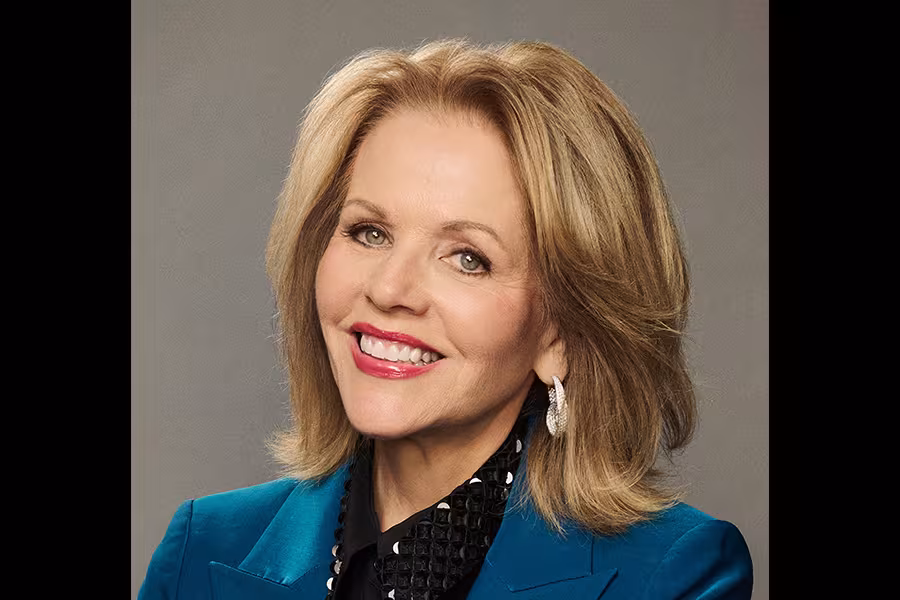 Portrait of Renée Fleming smiling at the camera against a gray background, wearing a teal blazer, black blouse, and silver earrings.
