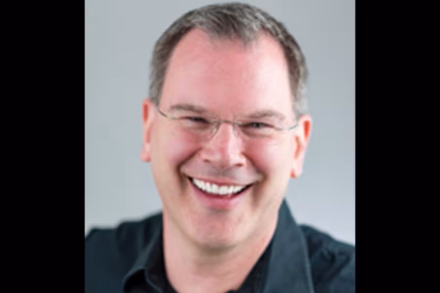 Headshot of Scott Temple smiling warmly at the camera, wearing glasses and a dark collared shirt against a light gray background.
